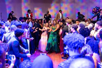 Students celebrating at the Black Excellence Gala in February of 2022. Two female students are dancing down an aisle created by other students. One is in a green formal dress, the other in light pink. All are cheering them on.