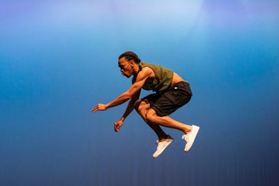 A member of Step Africa performs at a show on VT's campus in February of 2022. He is jumping in a crouched position, a blue screen filling all directions around him.