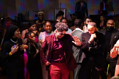 Young man in red velvet suit is centered in the photo, dancing with others behind him.