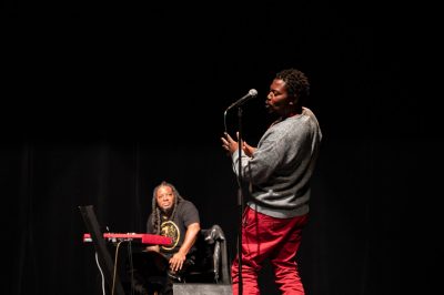Man stands at a microphone with his back slightly turned to the audience. He is clapping, dancing, and speaking into the microphone