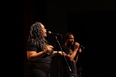 Woman from The Poetry Cafe singing into a microphone with a man out of focus giver her his attention in the distance.