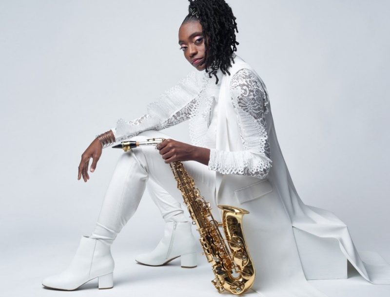 Lakecia Benjamin Image of Lakecia Benjamin dressed in all white with a saxophone to her side.
