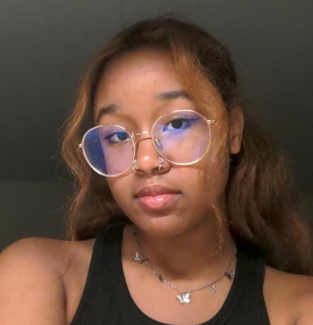 Taelor Baber, BCC graduate assistant. Closeup of a young Black woman with round, gold, wire-rimmed glasses looks into the camera. She is wearing a black shirt and a silver necklace with butterfly charms.