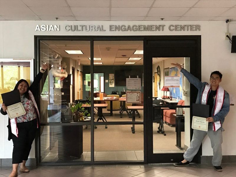Two students holding diplomas and wearing stoles in front of the ACEC entrance